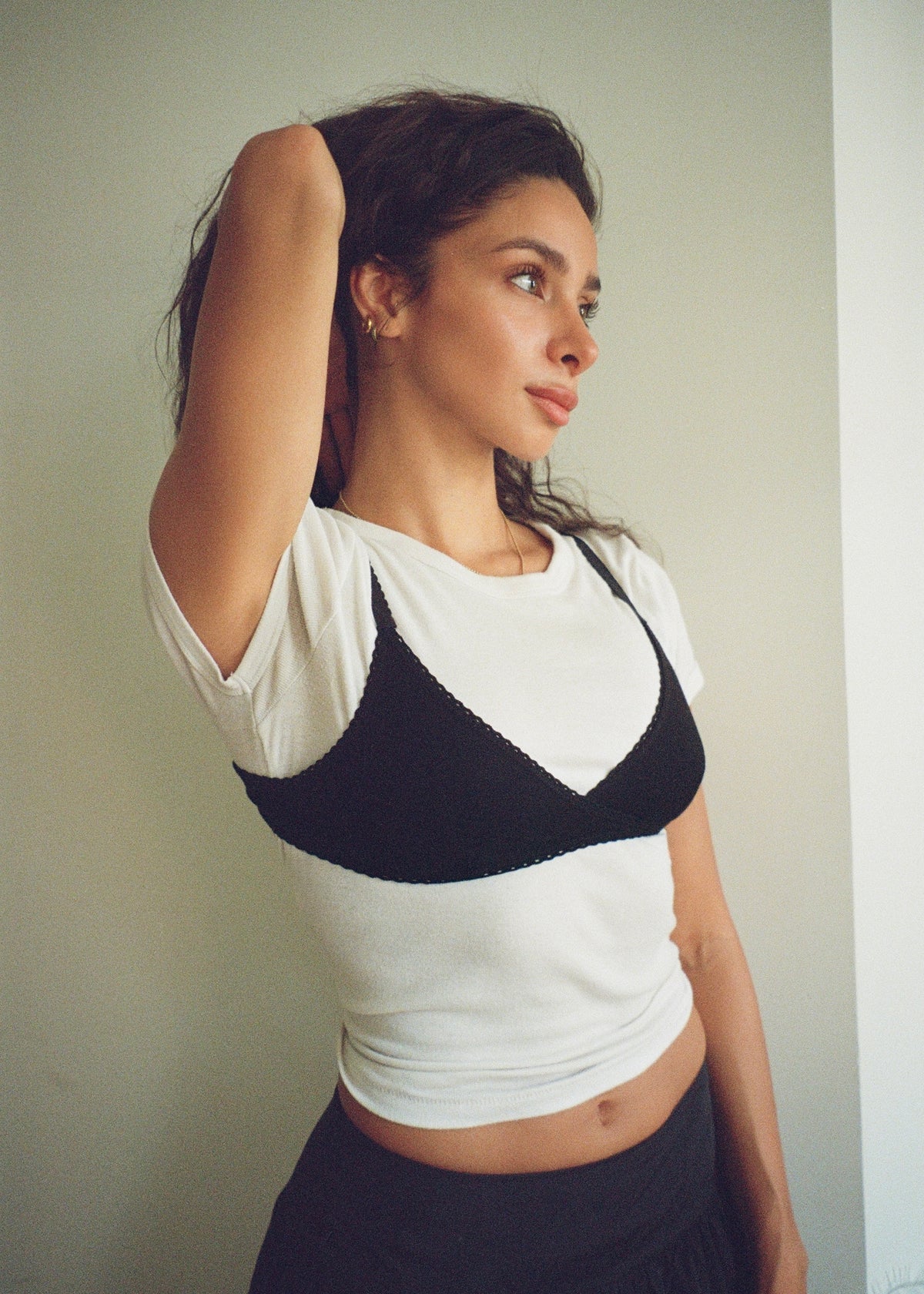 A woman wearing a white short-sleeve crop top with a black lunetti bra, paired with a dark skirt, standing against a plain light gray wall.