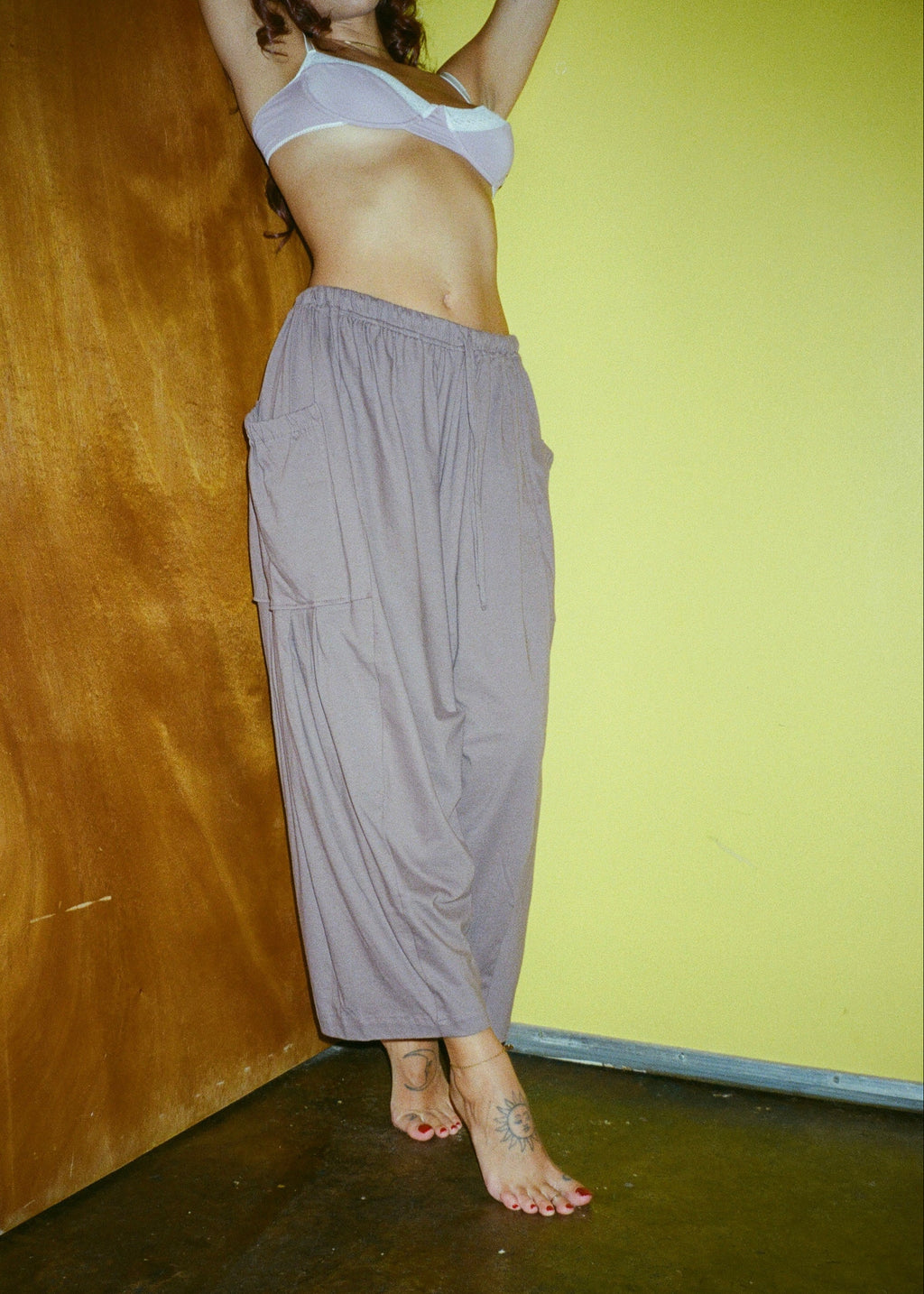 Model wearing the Mushroom Bailey Pant by Lunetti with a white bra, standing barefoot against a yellow wall and wooden door, showcasing the relaxed wide-leg silhouette.