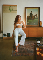 Model wearing the Stone Charlotte Bra by Lunetti with light-wash jeans, seated on a chair in a warmly lit interior with a wooden console and framed artwork behind her.