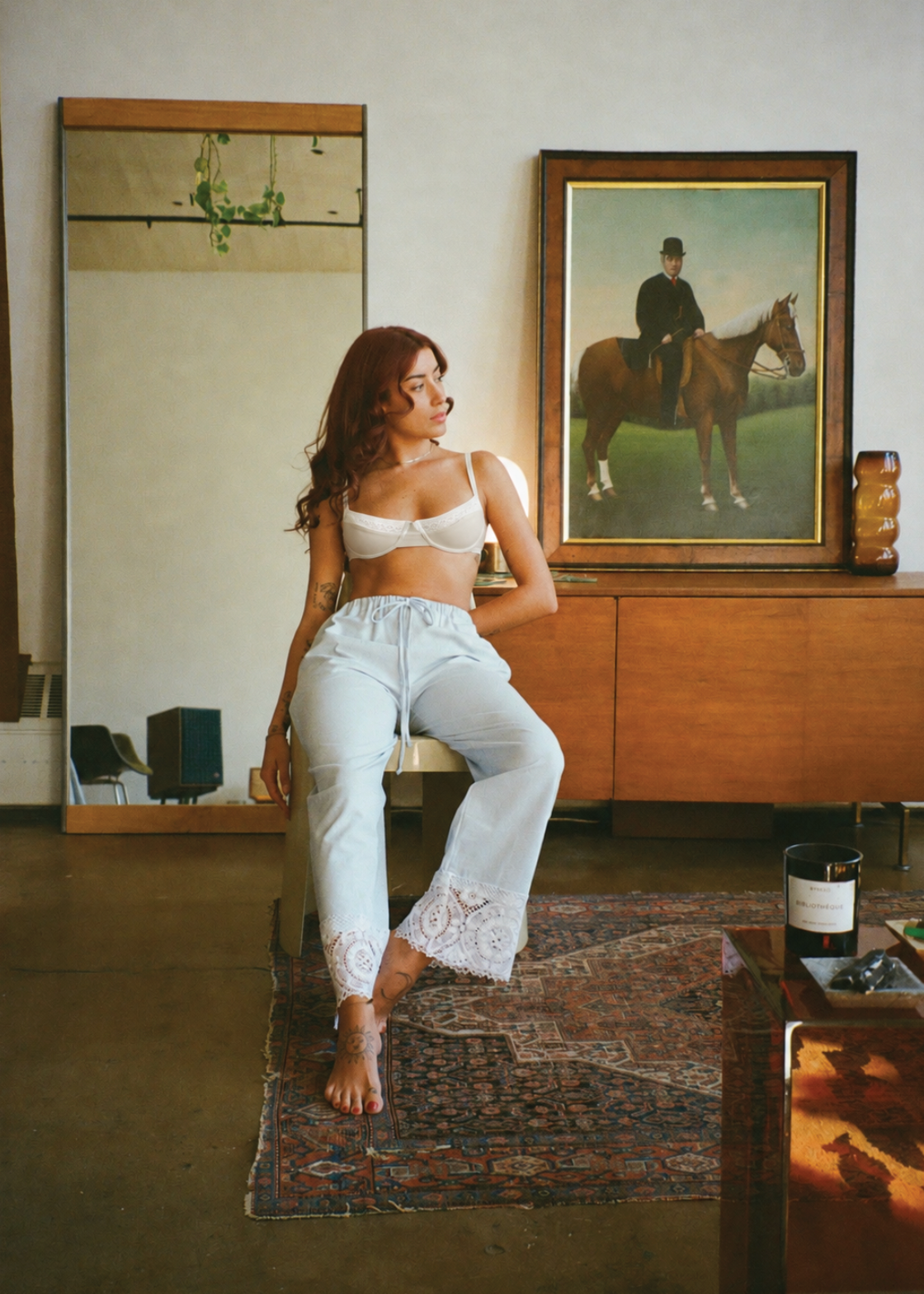Model wearing the Stone Charlotte Bra by Lunetti with light-wash jeans, seated on a chair in a warmly lit interior with a wooden console and framed artwork behind her.