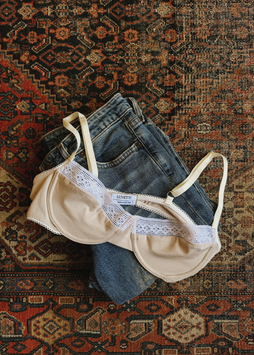 Stone Charlotte Bra by Lunetti styled with light-wash denim on a patterned vintage rug, showcasing the soft bamboo jersey, underwire shape, and delicate white lace trim.
