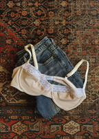 Stone Charlotte Bra by Lunetti styled with light-wash denim on a patterned vintage rug, showcasing the soft bamboo jersey, underwire shape, and delicate white lace trim.