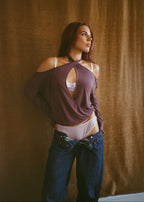 Model wearing the Amethyst Dahlia Top by Lunetti with low-rise blue jeans, posing against a brown curtain with the crisscross neckline draped off one shoulder and a white bralette visible underneath.