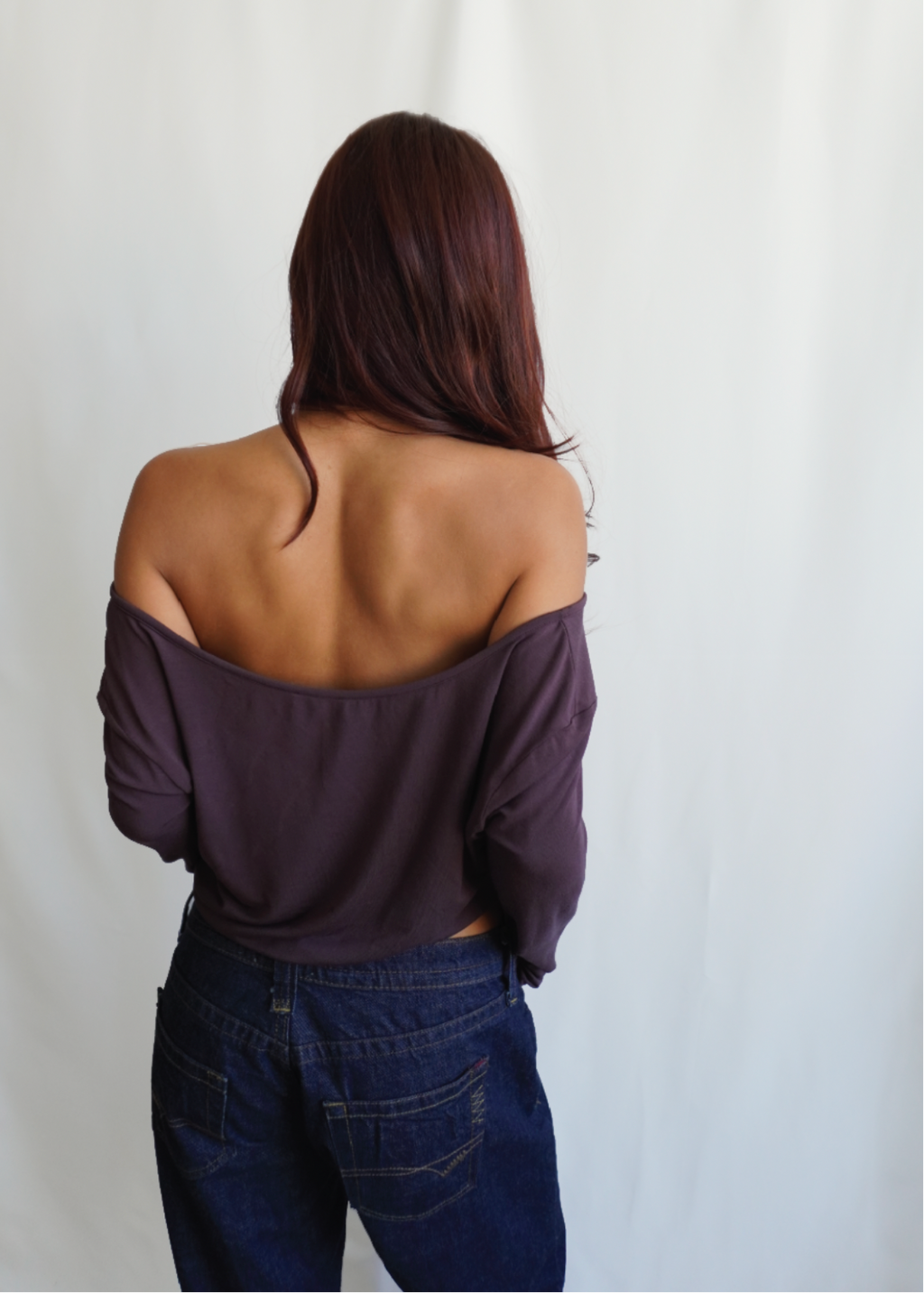 Rear view of a model wearing the Amethyst Dahlia Top by Lunetti with blue jeans, showcasing the wide off-the-shoulder neckline, softly draped back, and long sleeves.