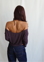 Rear view of a model wearing the Amethyst Dahlia Top by Lunetti with blue jeans, showcasing the wide off-the-shoulder neckline, softly draped back, and long sleeves.