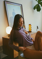 Model reclining on a bed wearing the Orchid Dahlia Top by Lunetti with matching underwear, showcasing the softly draped crisscross neckline and long sleeves in a warm, softly lit bedroom.