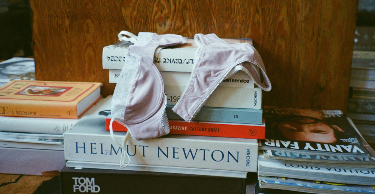 Lilac Charlotte Bra and matching thong by Lunetti draped over a stack of fashion photography books and magazines, showcasing the soft bamboo jersey fabric and delicate white lace trim.