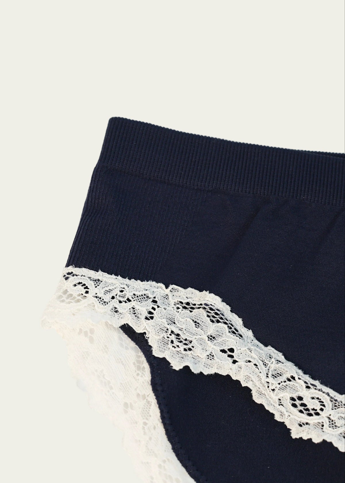Close-up of the Midnight Navy Luna Brief by Lunetti against an ivory background, showcasing the smooth navy fabric, waistband stitching, and scalloped ivory floral lace trim.