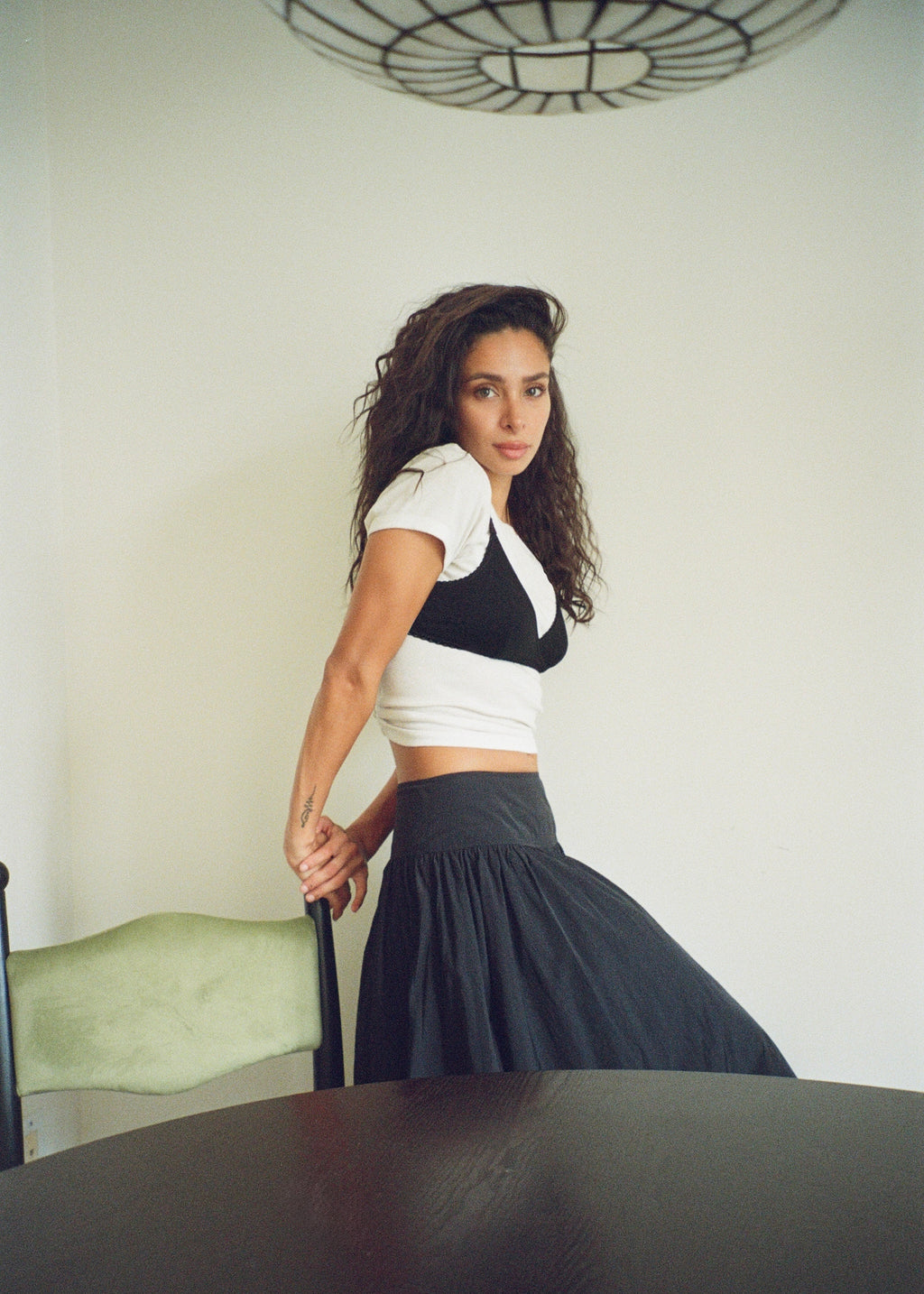 Model wearing the Black Bamboo Margot Bralette by Lunetti over a white T-shirt, paired with a dark skirt, posing sideways in a sunlit interior.