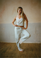 A woman modeling a light sage green lounge set consisting of a camisole top with white lace trim and matching sweatpants, standing in a rustic room with a textured plaster wall and wooden floor.