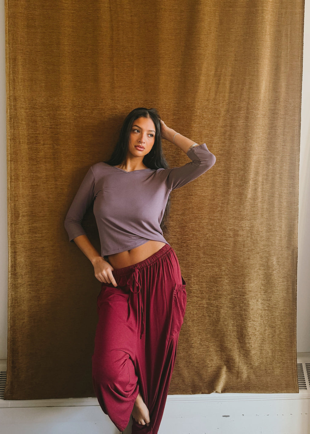 Model wearing the Amethyst Willow Top by Lunetti with burgundy pants, posing in front of a brown curtain, highlighting the fitted sleeves and soft bamboo fabric.