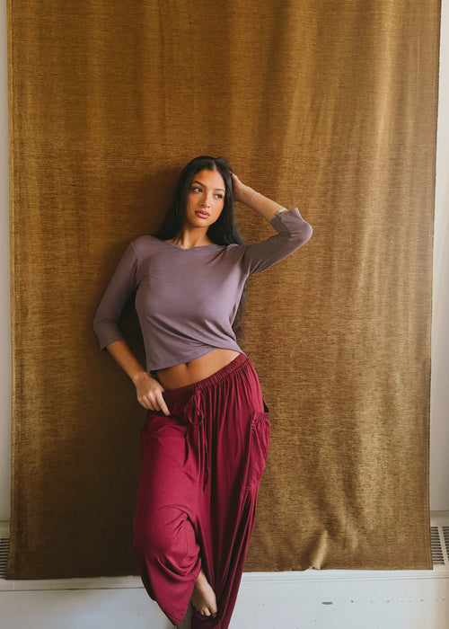 Model wearing the Amethyst Willow Top by Lunetti with burgundy pants, posing in front of a brown curtain, highlighting the fitted sleeves and soft bamboo fabric.