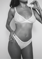 A black and white photo of a model wearing a matching white lace bra and panty set against a plain light gray background.