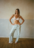 A woman stands barefoot in a studio with a textured beige and gray wall and wooden floor, wearing a white lace-trimmed bra and pale blue wide-leg pants with lace cuffs.