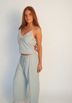 A woman modeling a light blue two-piece loungewear set consisting of a camisole top with white lace trim and matching wide-leg pants, set against a plain light background.