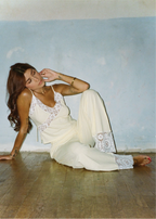 A woman reclining on a wooden floor against a light blue wall, wearing a cream-colored lace-trimmed camisole and matching wide-leg pants.