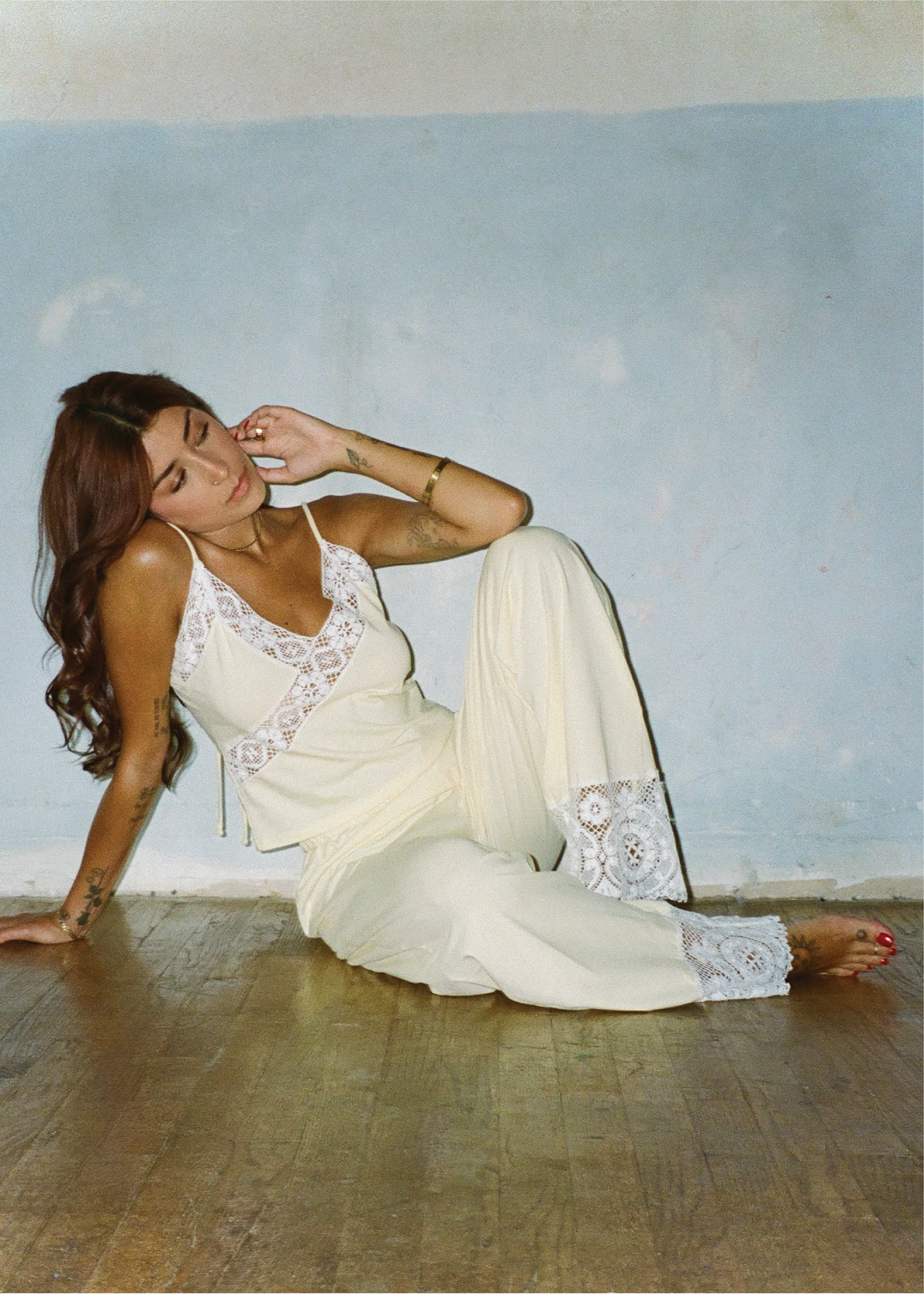 A woman reclining on a wooden floor against a light blue wall, wearing a cream-colored lace-trimmed camisole and matching wide-leg pants.