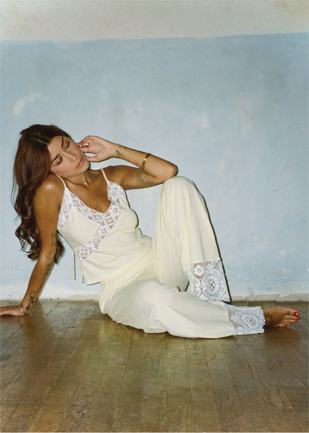 A woman reclining on a wooden floor against a light blue wall, wearing a cream-colored lace-trimmed camisole and matching wide-leg pants.