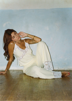A woman reclining on a wooden floor against a light blue wall, wearing a cream-colored lace-trimmed camisole and matching wide-leg pants.
