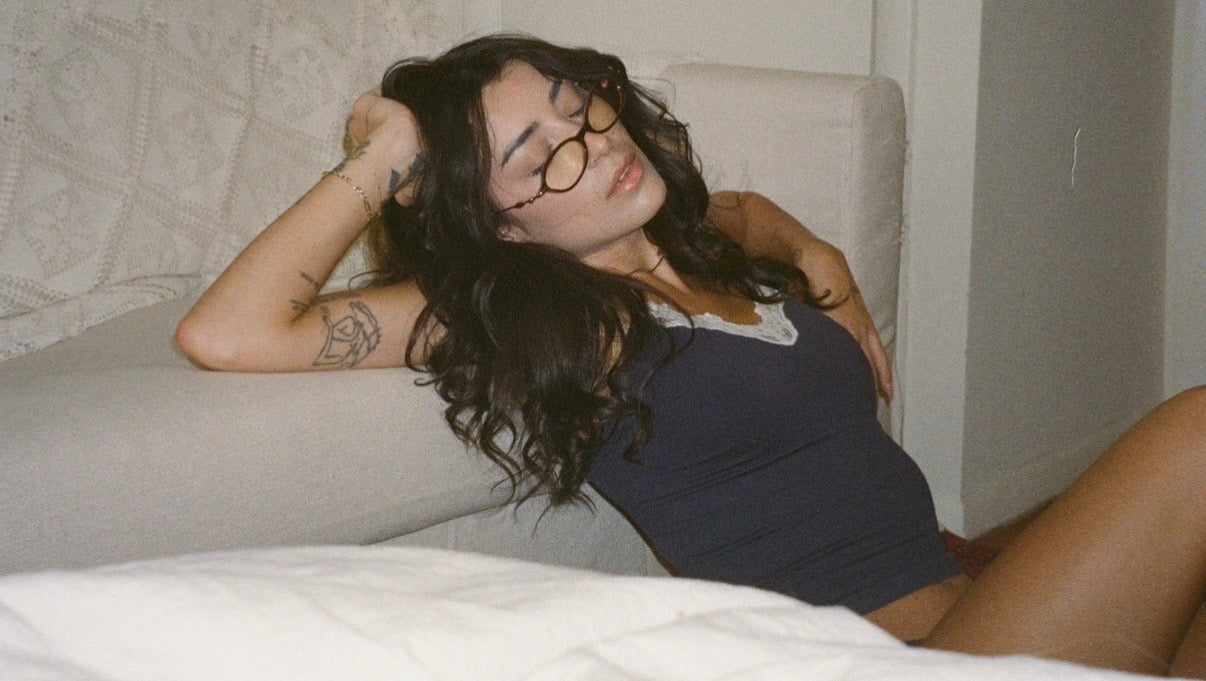 Woman reclining beside a bed wearing the Lunetti Luna Cami in midnight navy with white lace trim, photographed for the Lunetti About page.